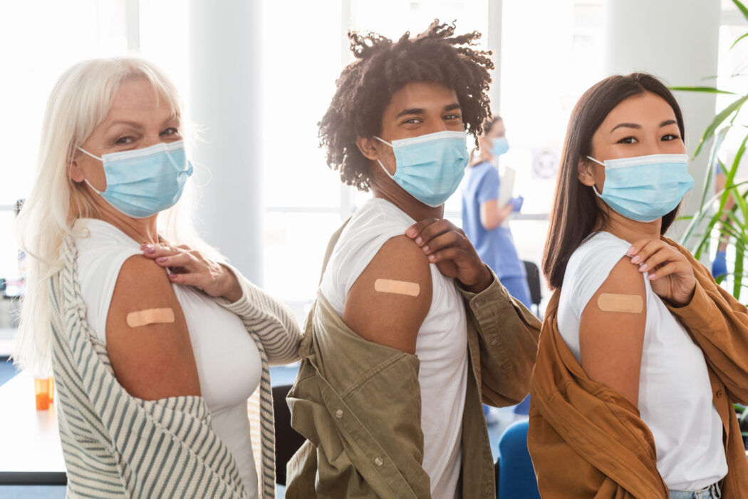 A few people wearing masks are showing off their bandaids after receiving a vaccine.