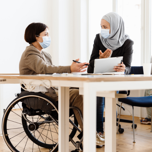 Two women in face masks talking at a table.