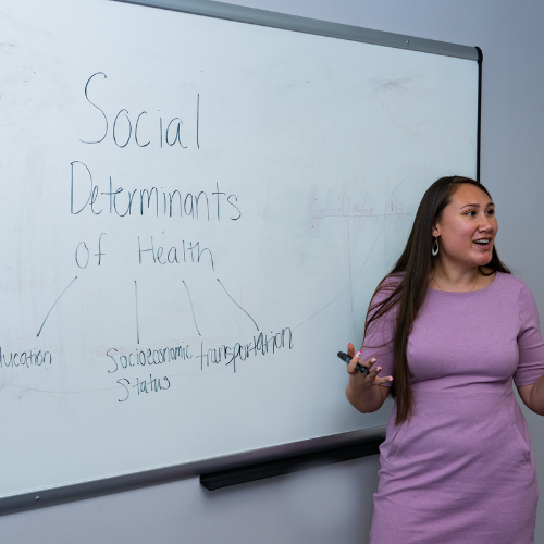 Woman standing in front a whiteboard teaching about the social determinants of health.