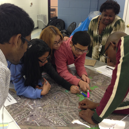 A group of teens and adults are gathered around a map with pens for a community workshop activity.