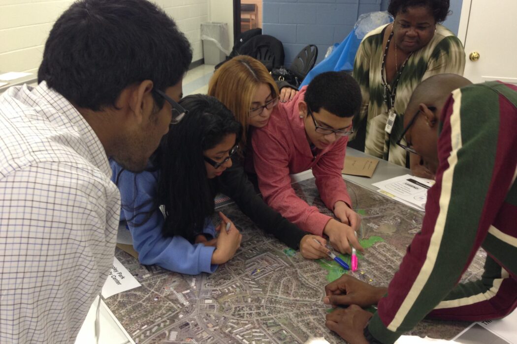 A group of teens and adults are gathered around a map with pens for a community workshop activity.