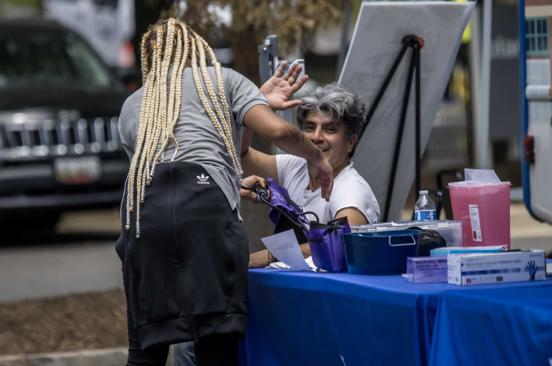 A woman reading a man's blood pressure at a tabling event. The woman's back is turned while the man is waving and smiling at the camera.