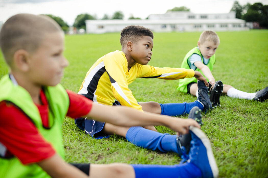Three boys wearing soccer gear are stretching on the field.