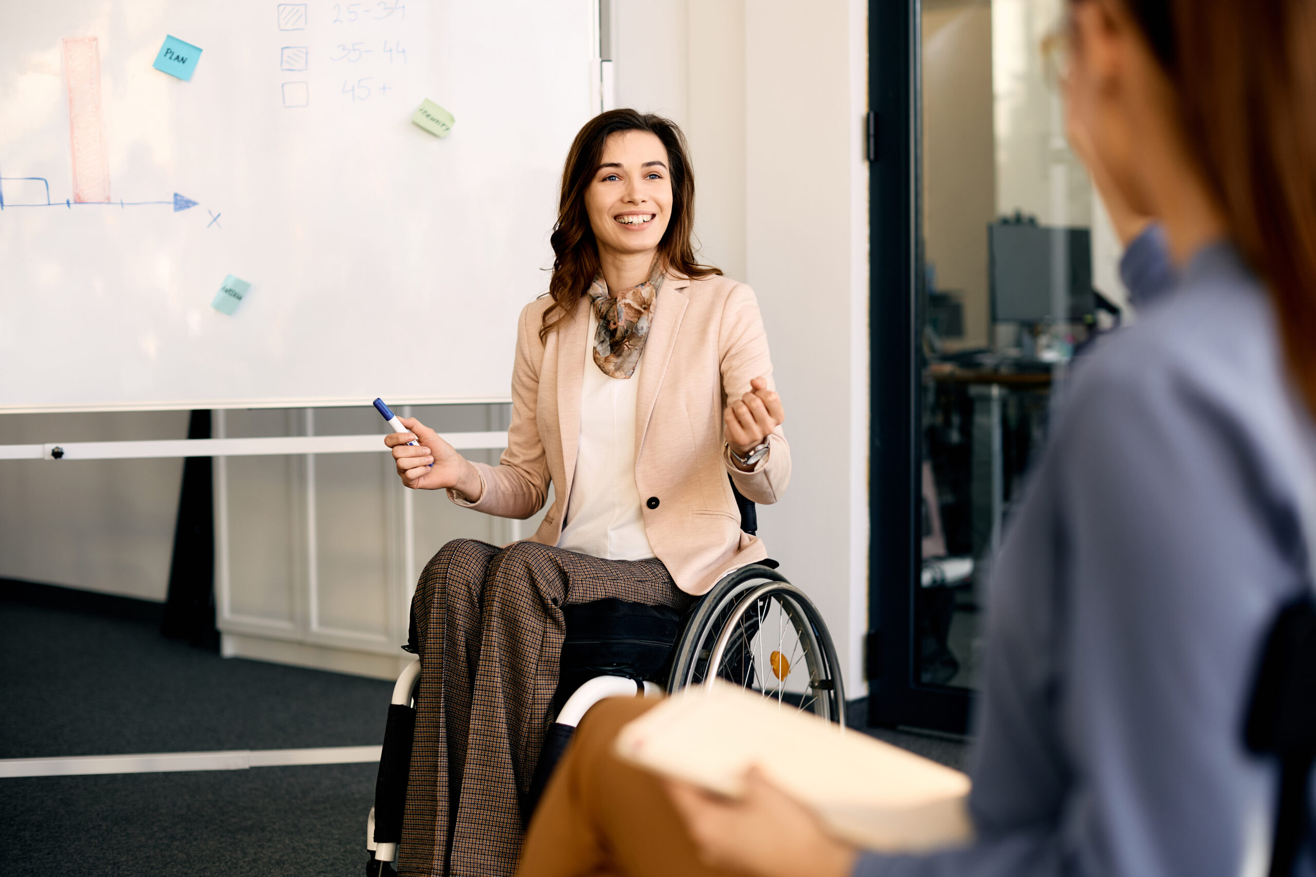 Happy businesswoman in wheelchair presenting new ideas on whiteboard to colleagues in the office.