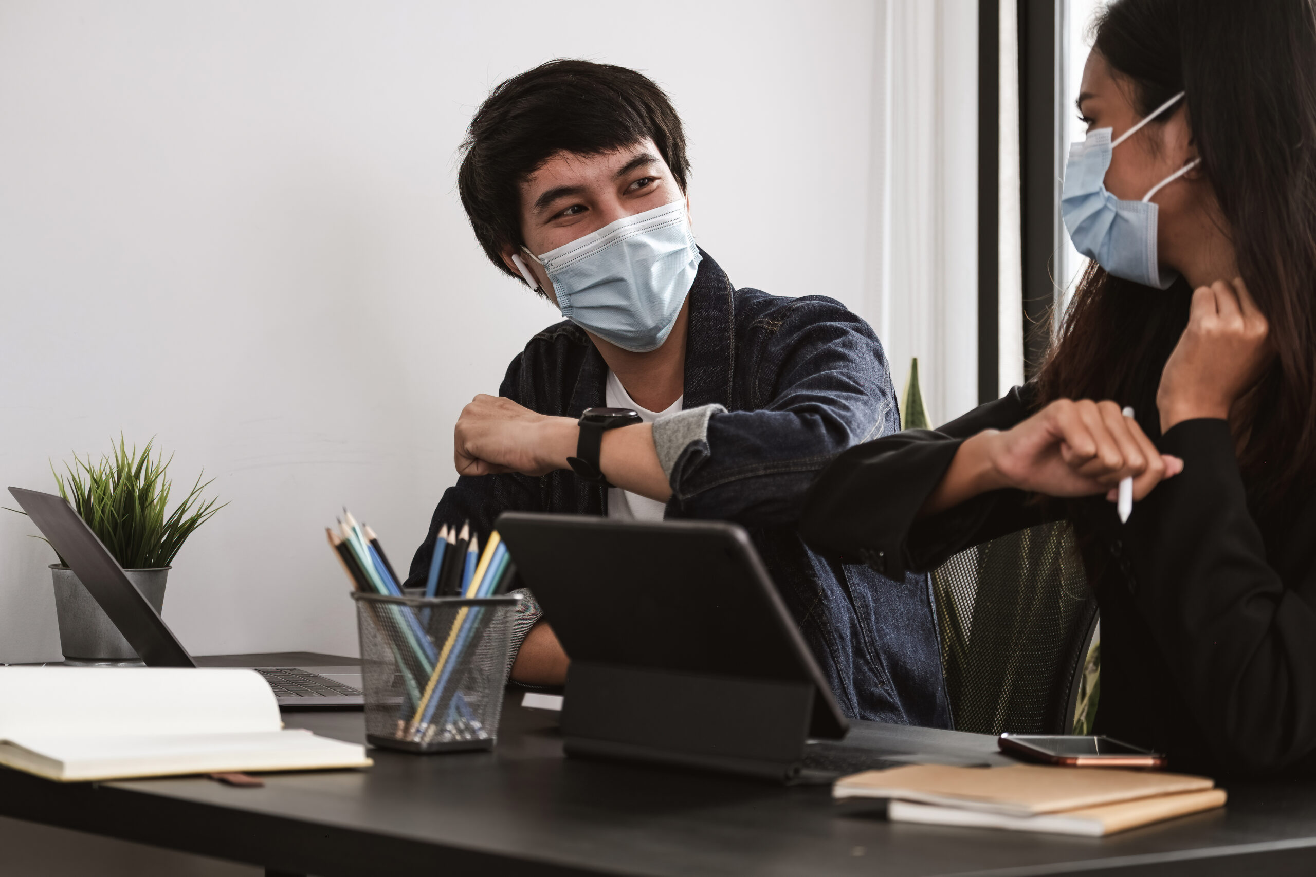 Two young diverse business colleagues wearing face protective masks bumping elbows, greeting each other while working during covid 19 quarantine